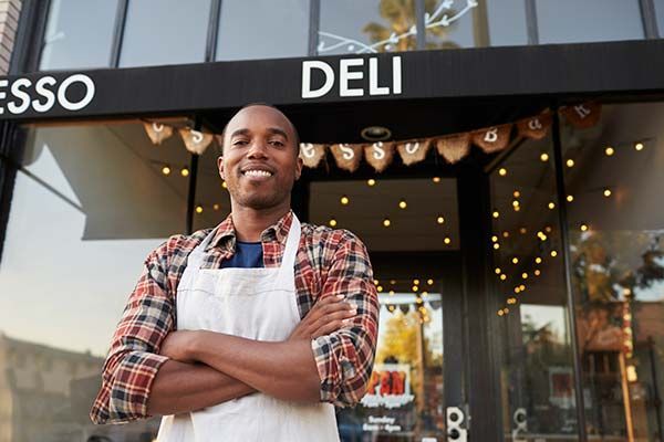Man in apron standing with arms crossed in front of deli, smiling. Exterior shot with awning.