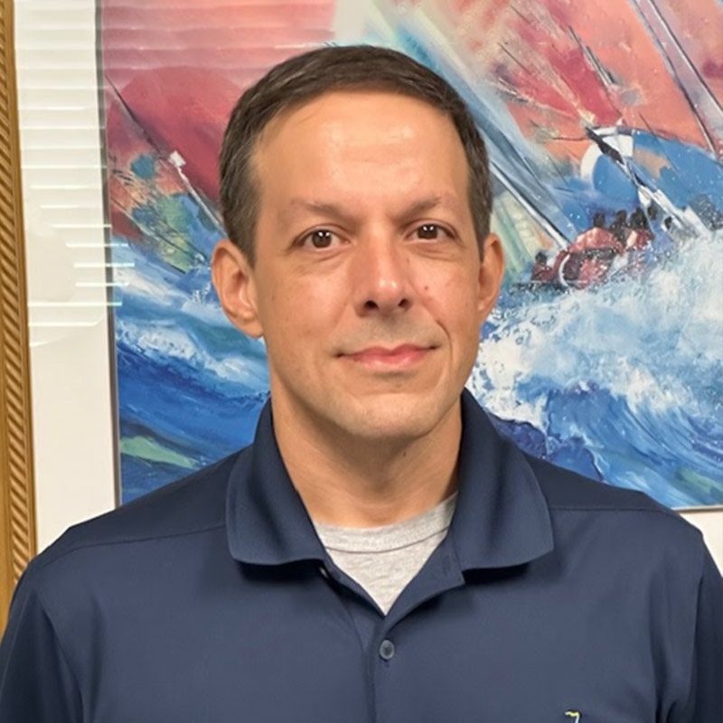 Man in navy polo shirt, gray undershirt, looking at the camera. Painting of sailboats in background.