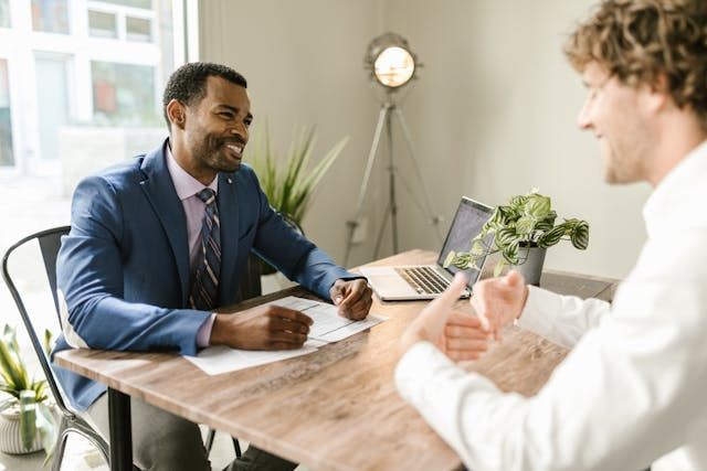 Two men smiling, engaged in conversation at a wooden table with a laptop, documents, and a plant.