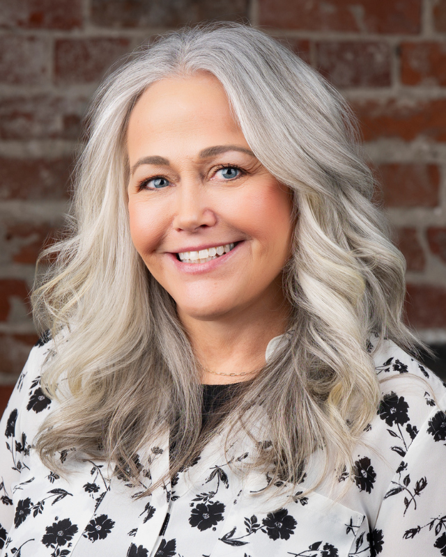A headshot of a smiling woman with white hair against a brick wall, and wearing a black and white patterned blouse