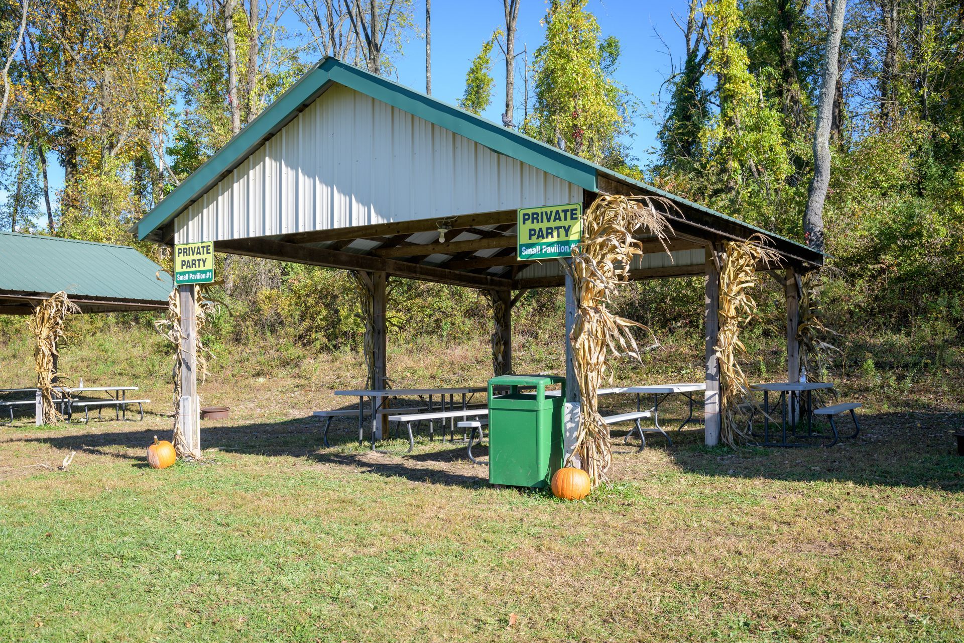 Picnic shelter in an outdoor setting, featuring a green roof, picnic tables, and a green trash can