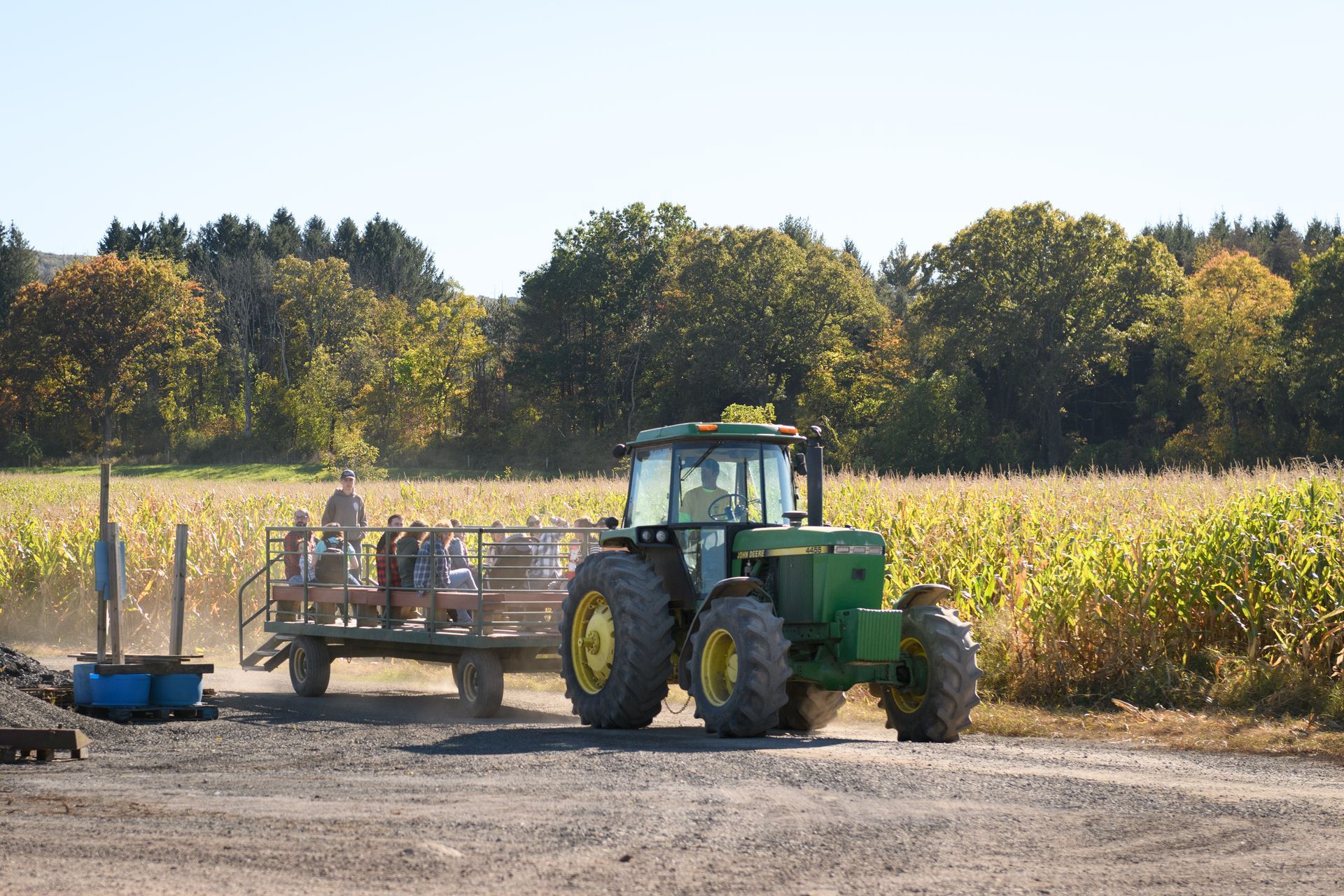A John Deere tractor pulls a wagon of people through a field. Sunny, fall day; corn stalks and trees visible.