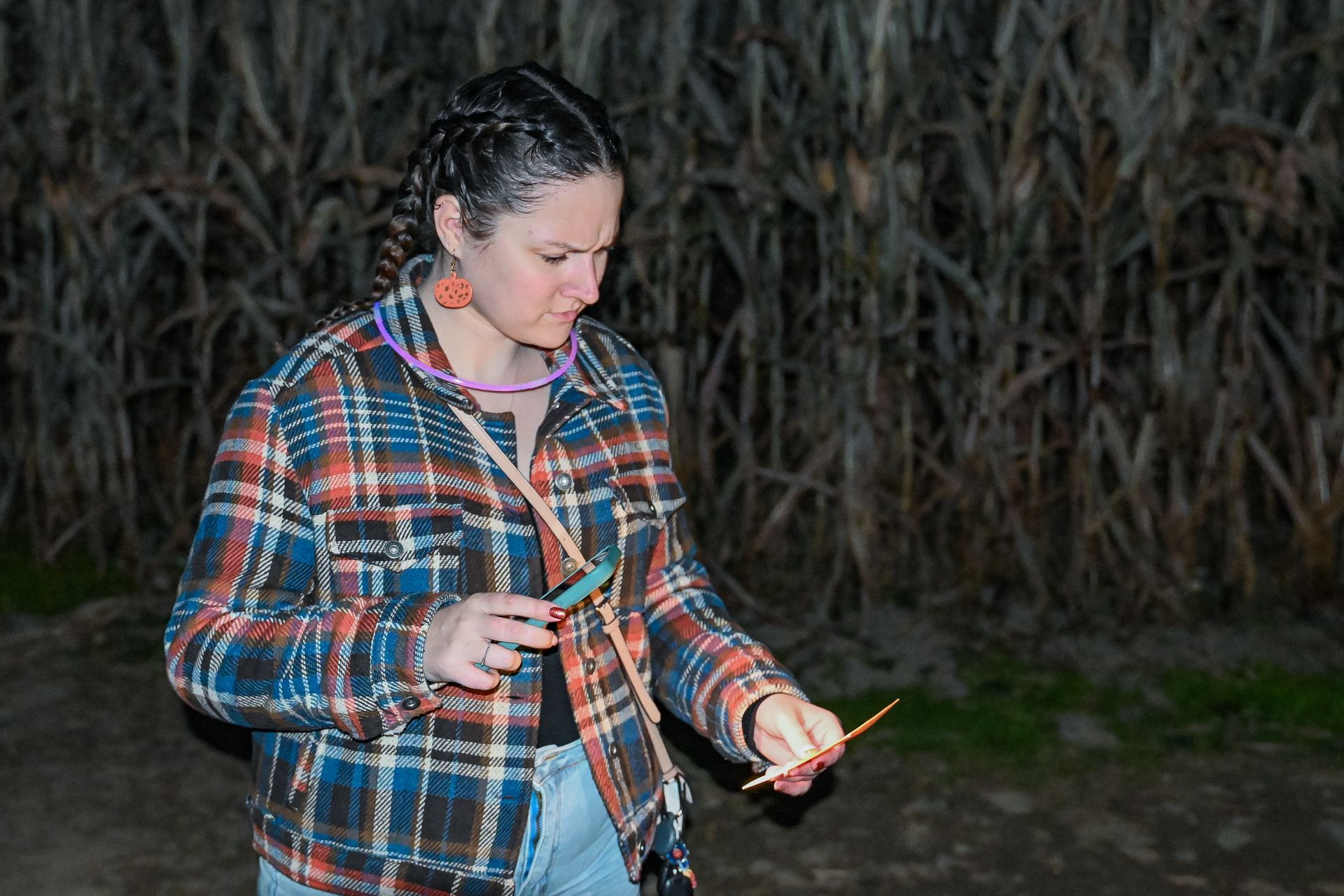 Woman in plaid shirt holding the map in a corn maze at night, looking down with a focused expression.