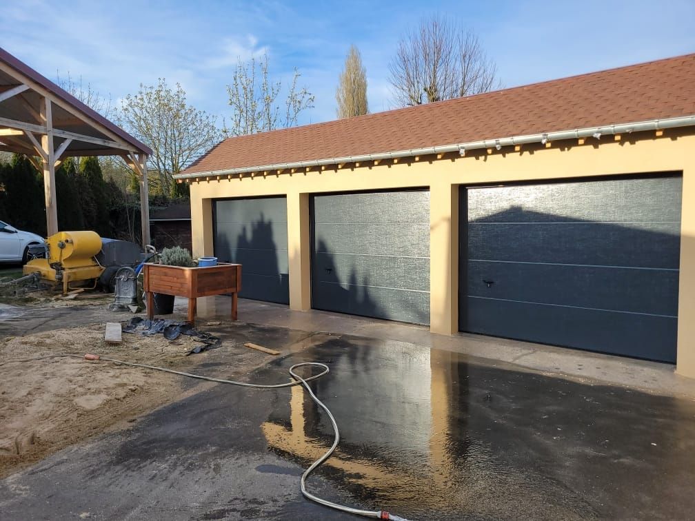 Garage pour trois voitures avec portes grises, façade beige et toit de tuiles rouges, par une journée ensoleillée.