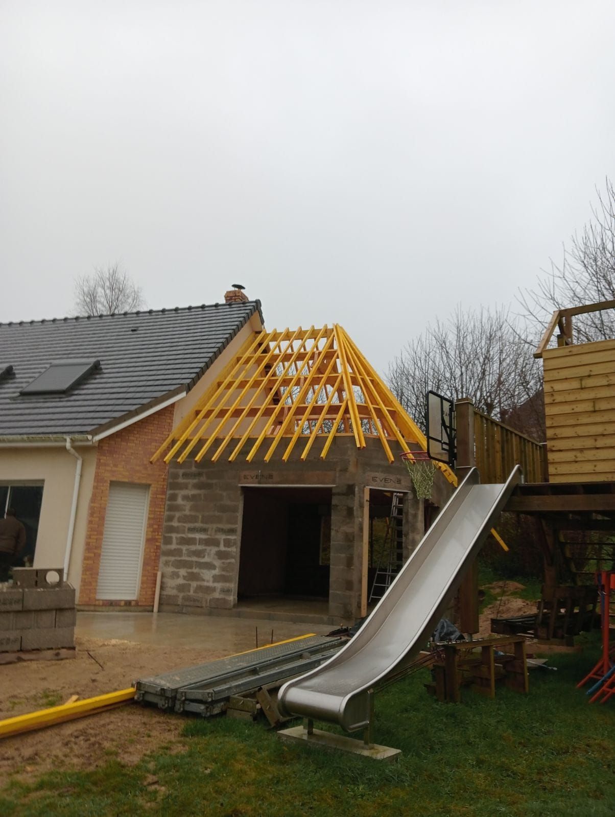 Maison en construction avec charpente apparente, toboggan et terrasse dans le jardin.