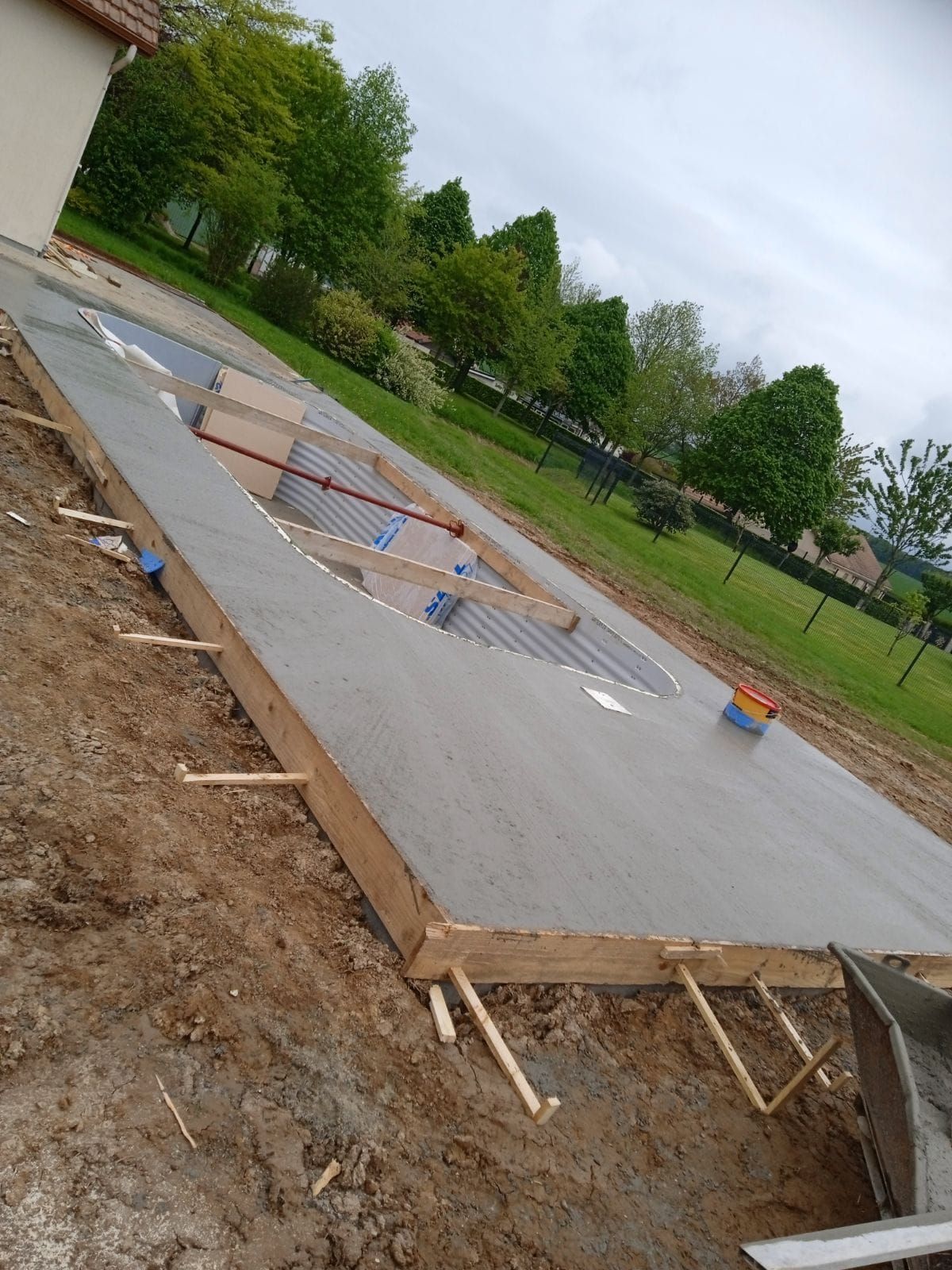 Dalle de béton en cours de réalisation, avec coffrage en bois et découpe. Extérieur, sol en terre battue, ciel couvert.