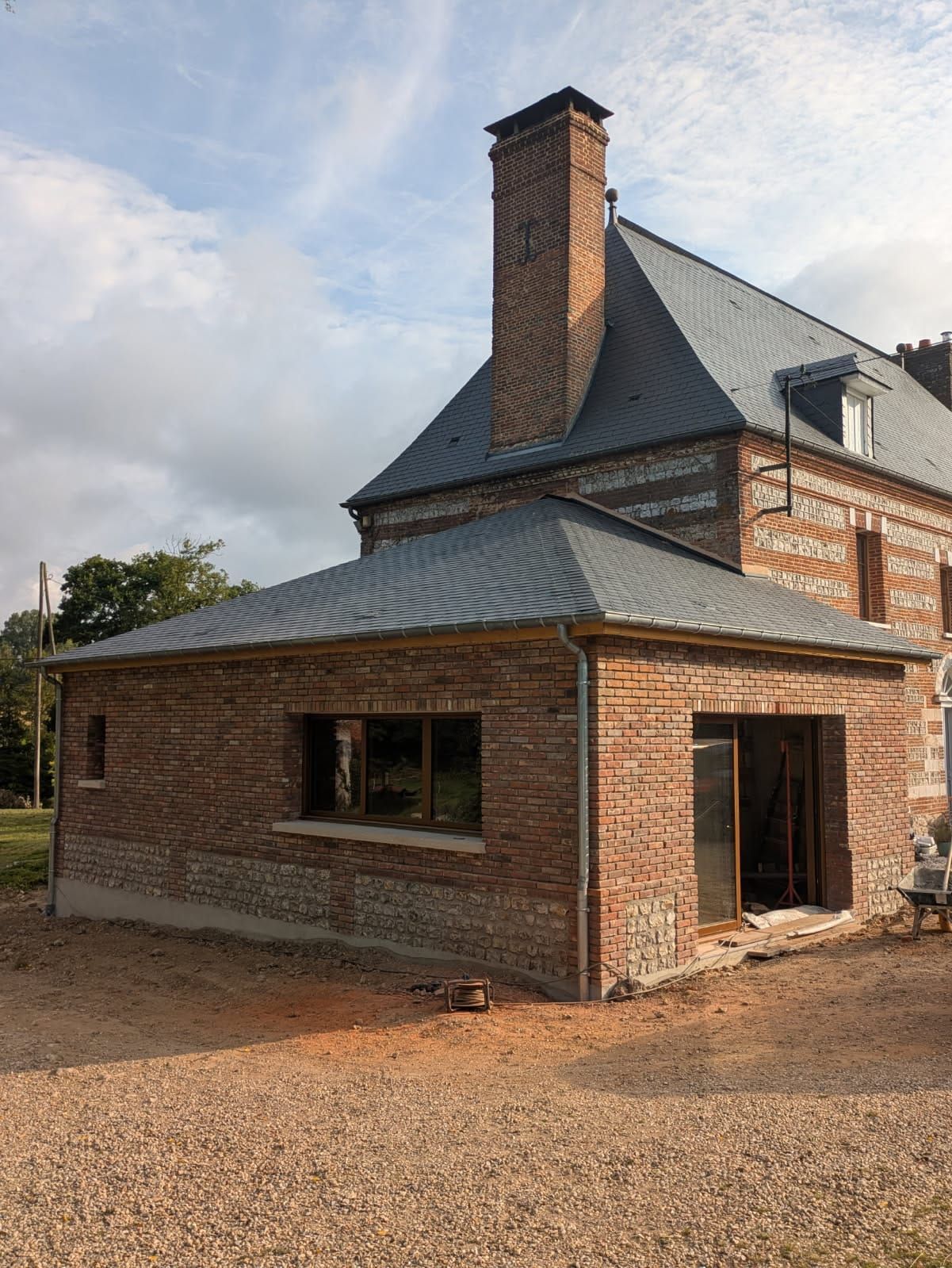 Bâtiment en briques avec une nouvelle extension en briques, une cheminée et un toit sombre sous un ciel nuageux.