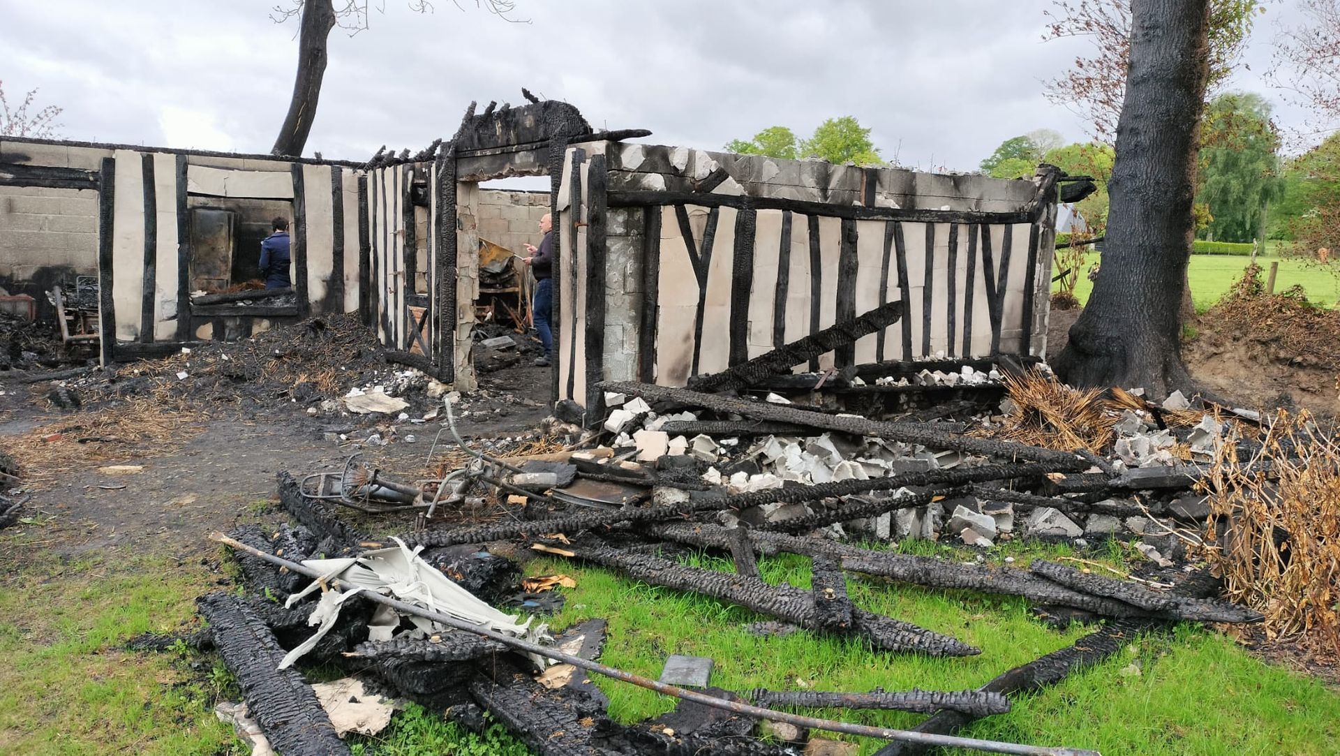 Vestiges d'un bâtiment à ossature bois incendié, avec du bois carbonisé et des débris sur l'herbe.