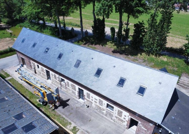 Bâtiment long et bas, au toit sombre et percé de plusieurs puits de lumière, en milieu rural.
