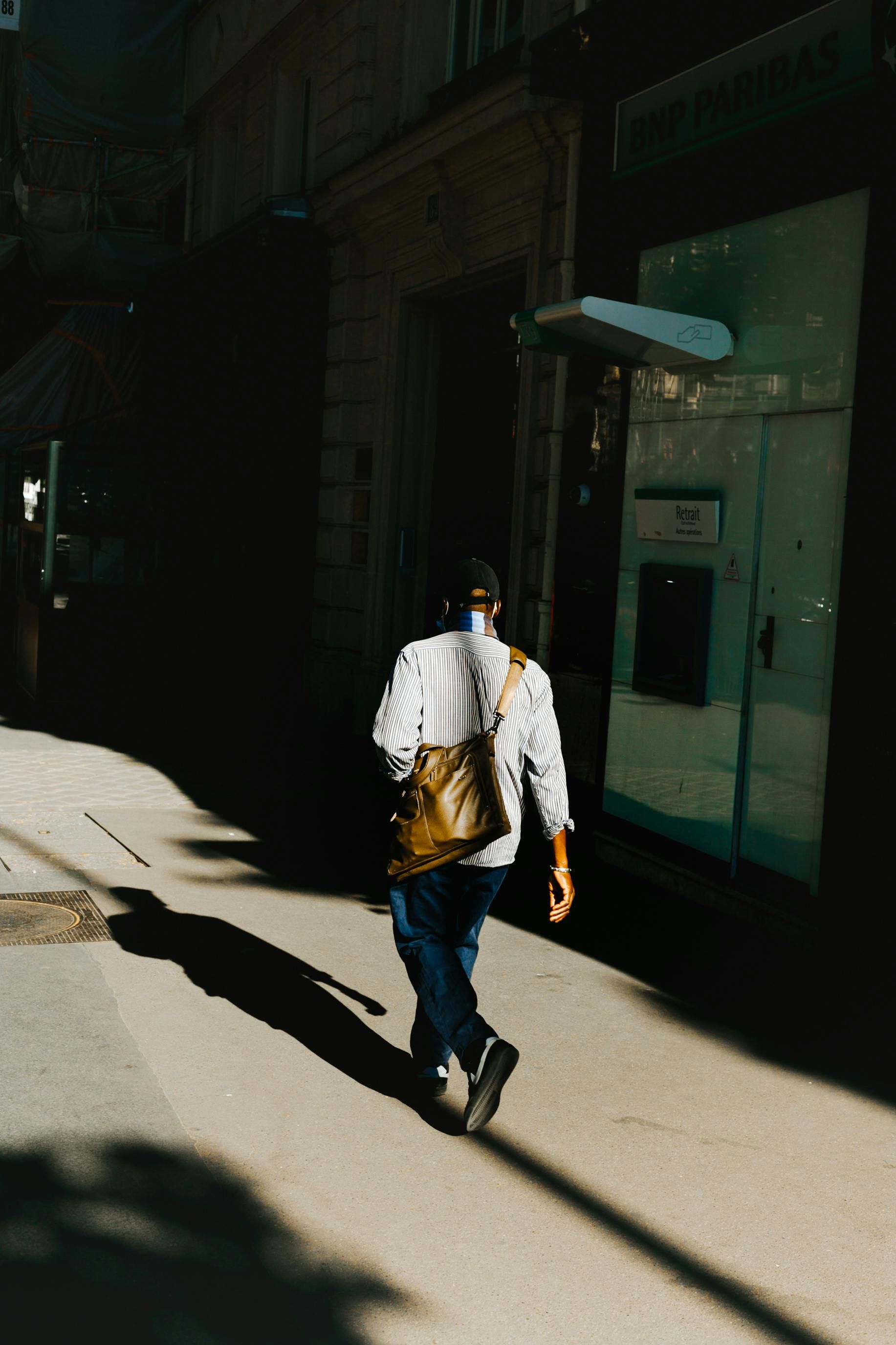 Une personne vêtue d'une chemise à rayures et d'un jean foncé marche sur un trottoir ensoleillé