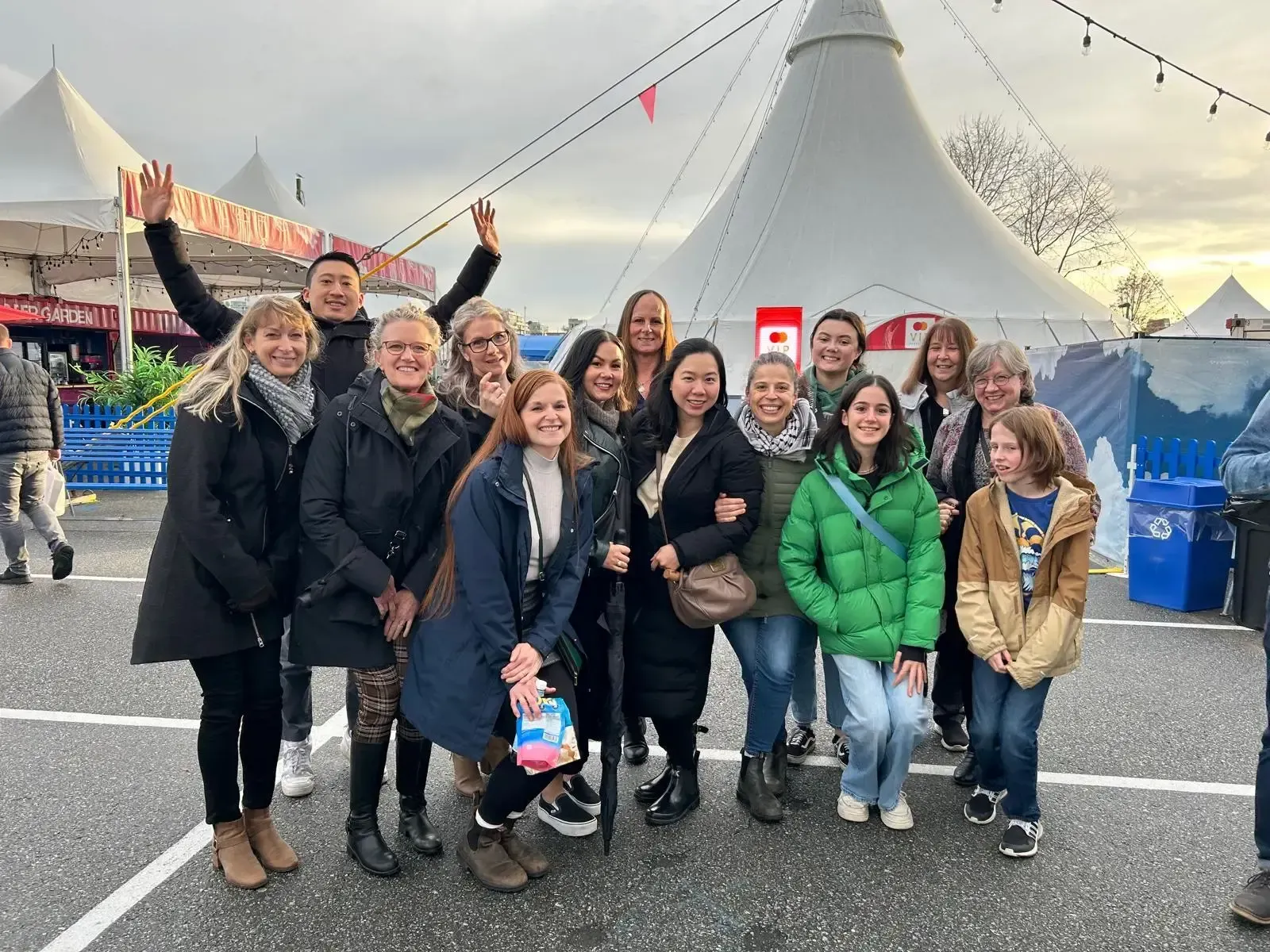 A large group poses for a photo outside at a festival with white tents in the background, many smiling and waving.