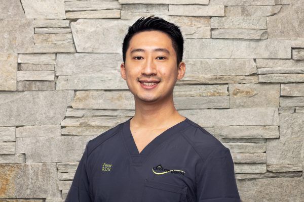 A professional portrait of a person in dark blue medical scrubs smiling in front of a gray stone wall.