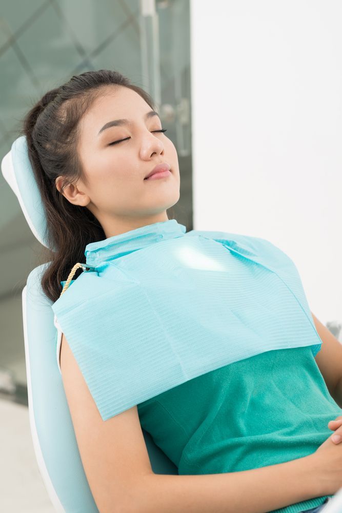 A person with eyes closed sits in a dental chair wearing a teal patient bib.