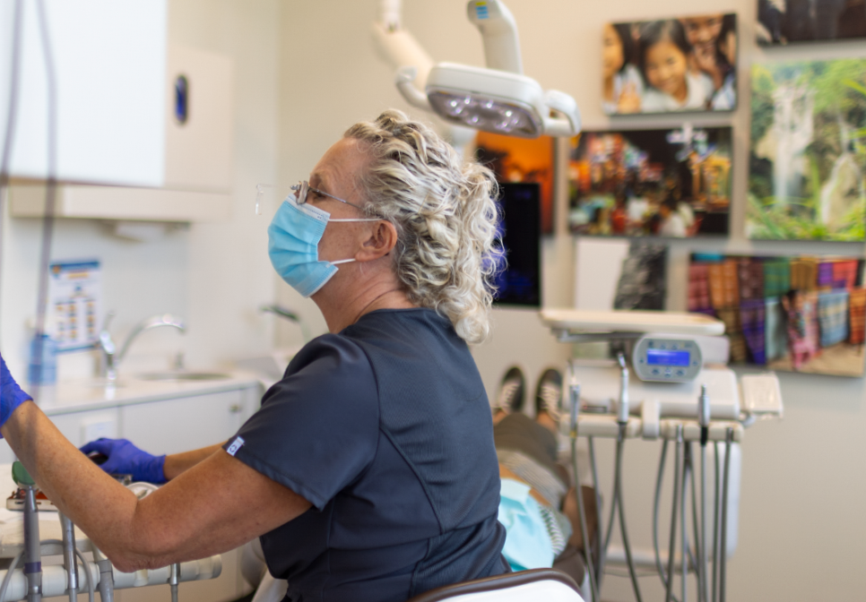 A professional in a mask and blue scrubs works at a dental station in a clinic filled with wall photos.