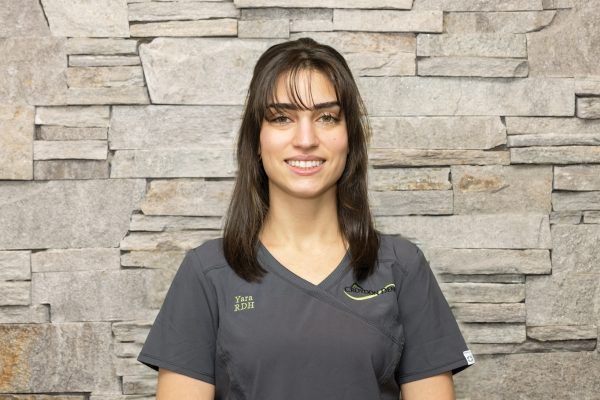 A person with dark hair and a light smile wearing a dark gray v-neck scrub top against a stone wall background.
