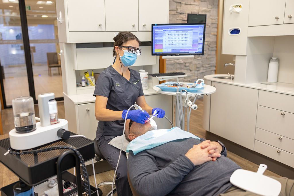 A dental professional in scrubs and a mask provides treatment to a patient in a chair inside a clinic.