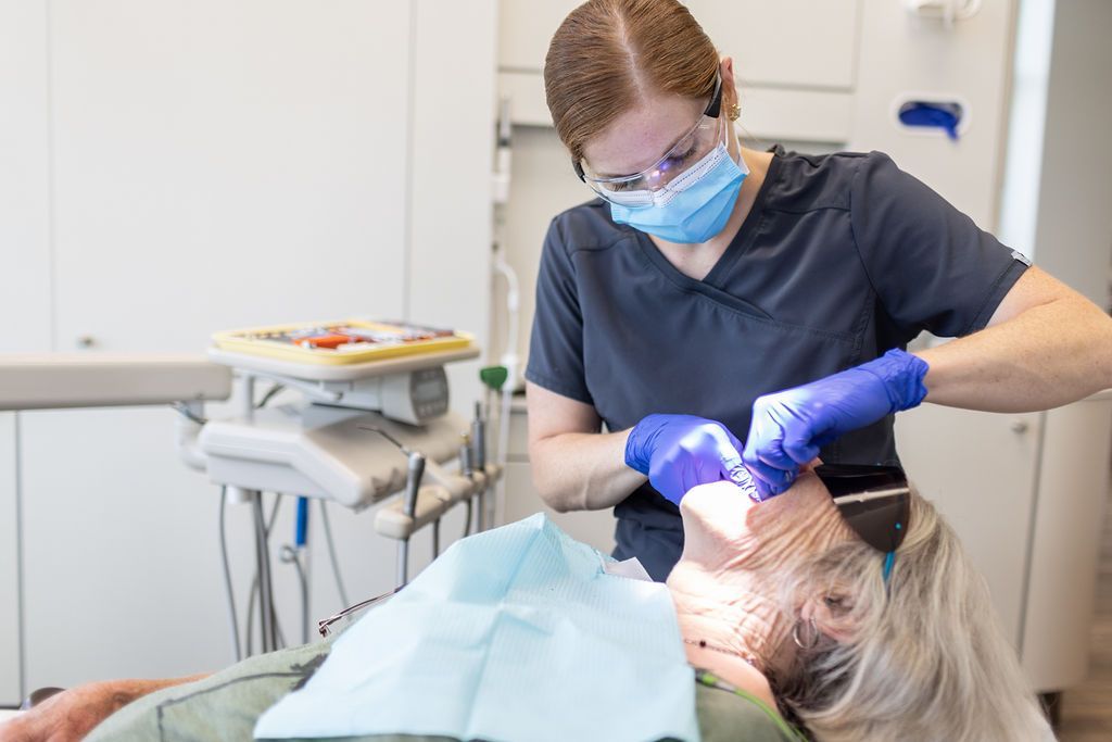 A dentist in blue scrubs and a face mask examines a patient lying in a dental chair.