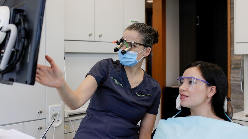 A dental professional wearing surgical loupes points to a screen while explaining treatment to a patient in a clinic.