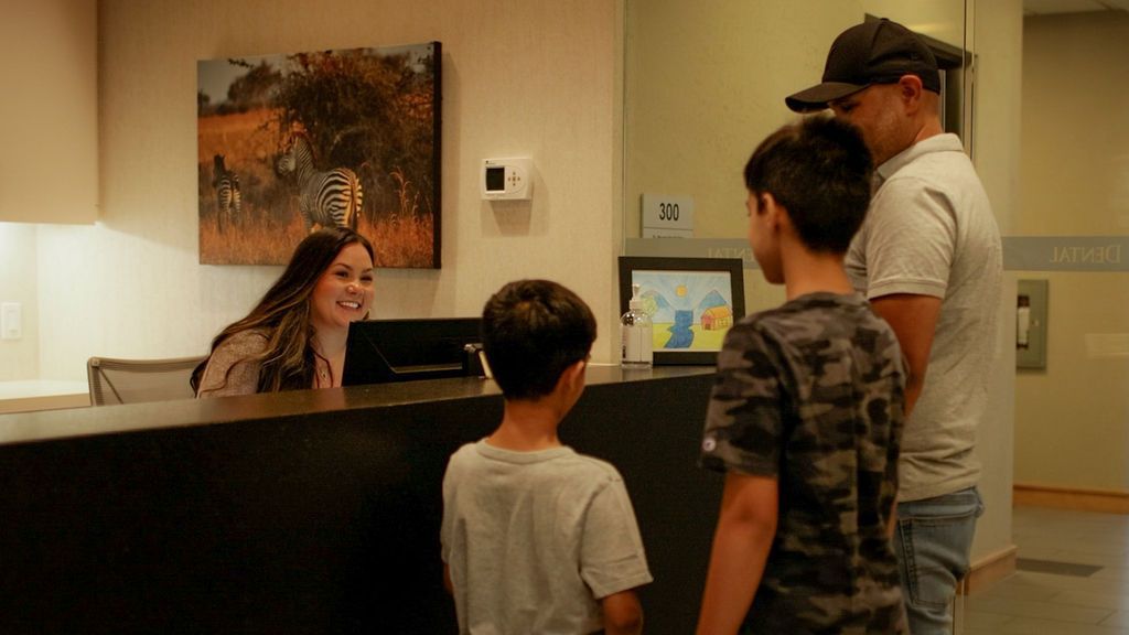 A staff member smiles at a family of three standing at a front desk in a brightly lit reception area.