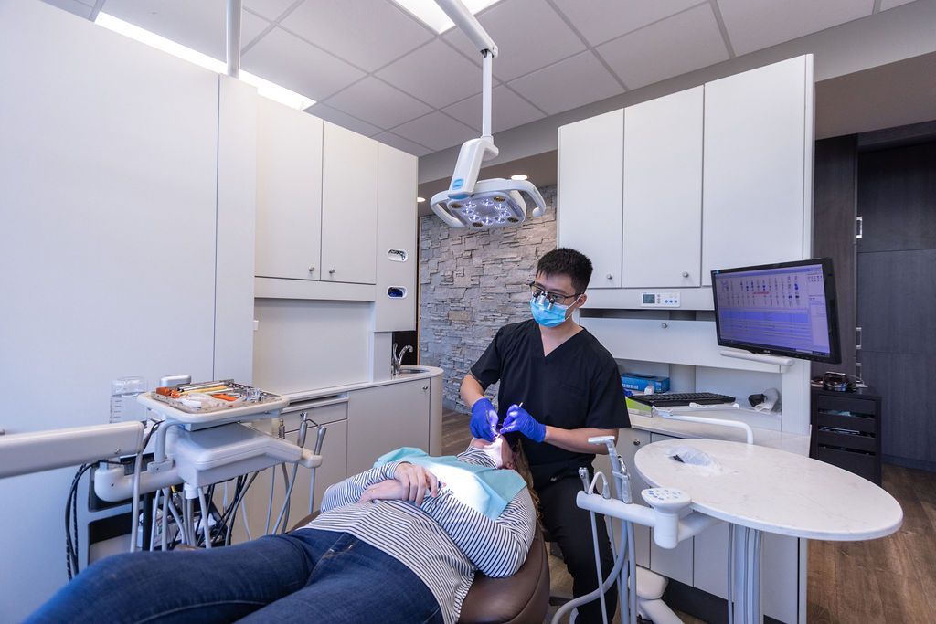 A dentist wearing a mask and blue gloves examines a patient lying in a dental chair in a clinical office setting.
