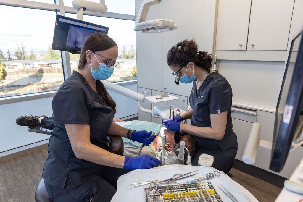 Two dental professionals in scrubs and masks perform a procedure on a patient in a bright dental office.