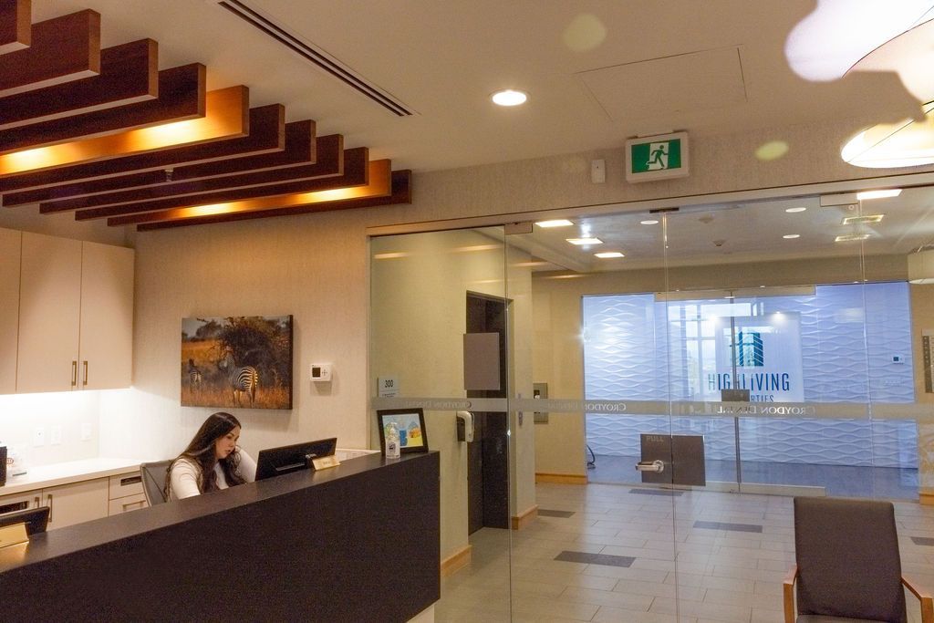 A receptionist sits at a dark desk in a modern, well-lit office lobby with wooden ceiling slats and a glass entryway.