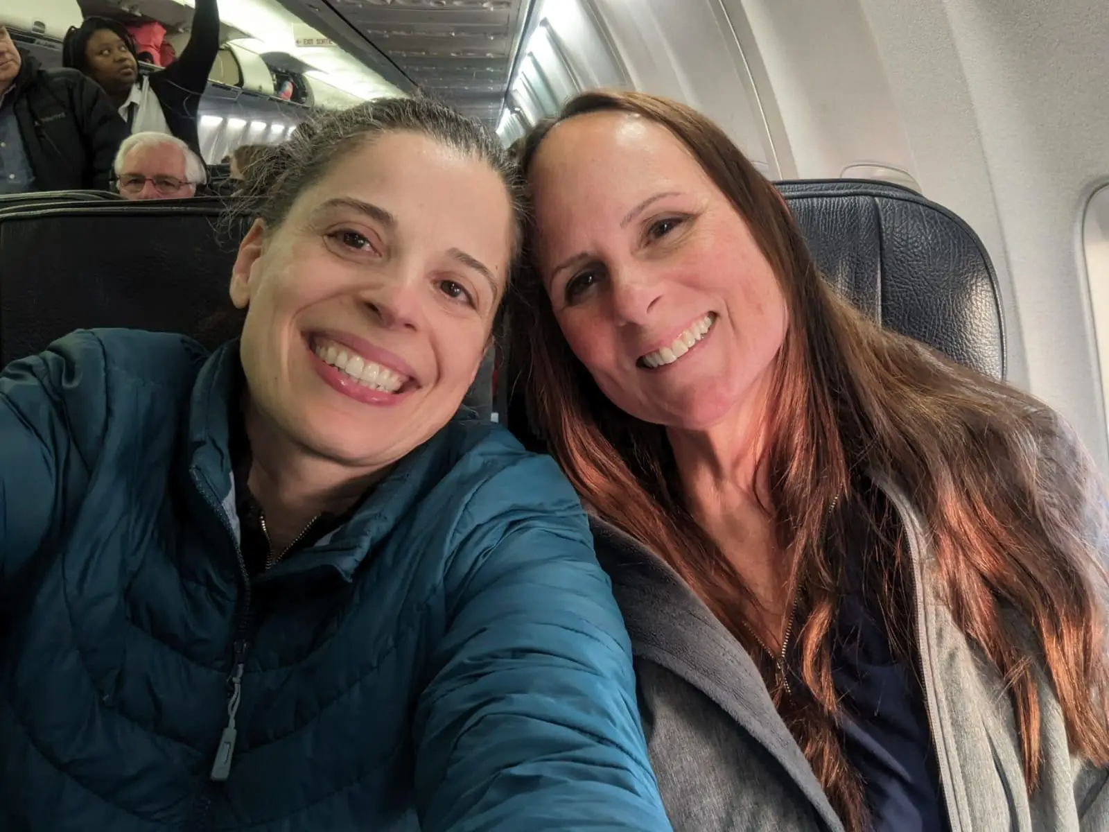 Two smiling people pose for a selfie while sitting in airplane seats.