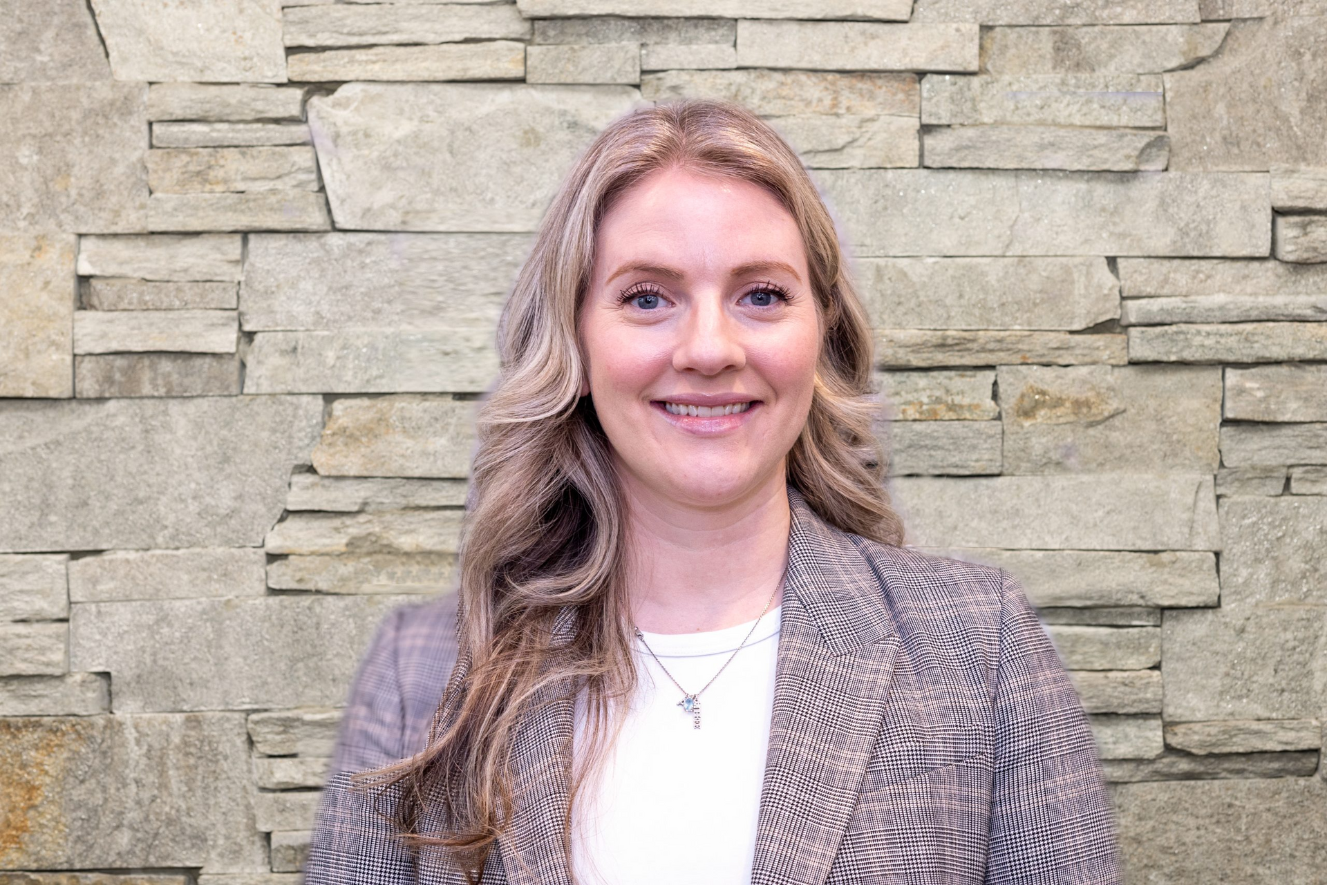 A smiling person with wavy hair wearing a plaid blazer over a white shirt, positioned against a textured stone wall.