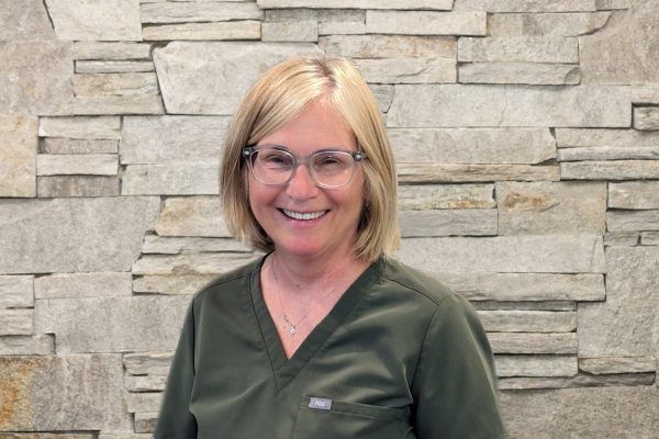 A smiling person wearing olive green medical scrubs and glasses, standing in front of a textured stone wall.