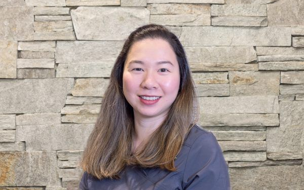 A person with long brown hair smiling at the camera, wearing a dark gray top against a textured stone wall background.