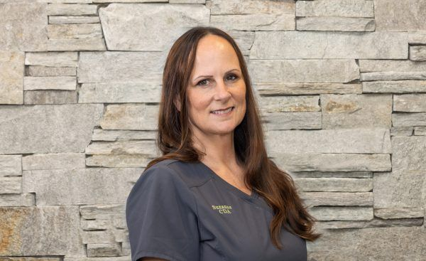 A person with long brown hair smiling softly, wearing a grey scrub top, posing against a textured stone wall.