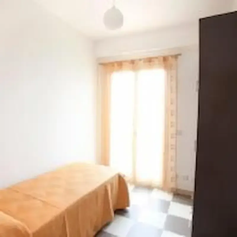 Small bright bedroom with a single bed, checkered floor, and sunlight through a window with curtains