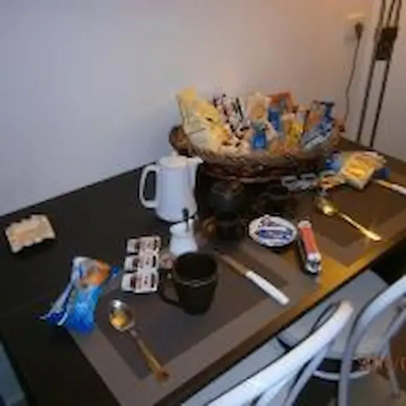 Cluttered dining table with snacks, mugs, utensils, and a large bowl in a plain room.