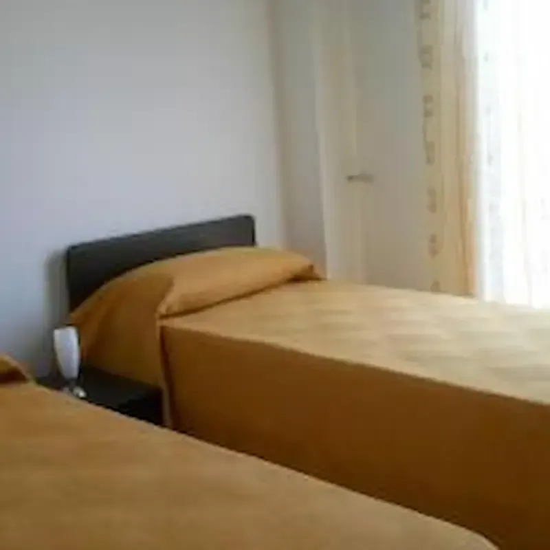 Small bedroom with two single beds made up in tan bedding beside a window and curtain.