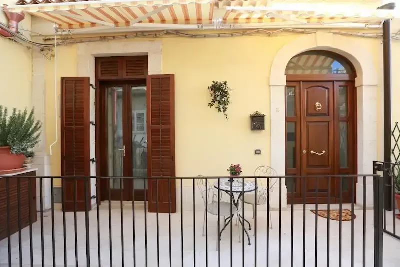 Shaded courtyard terrace with open wooden shutters, a wooden door, and a small table and chair behind a black railing