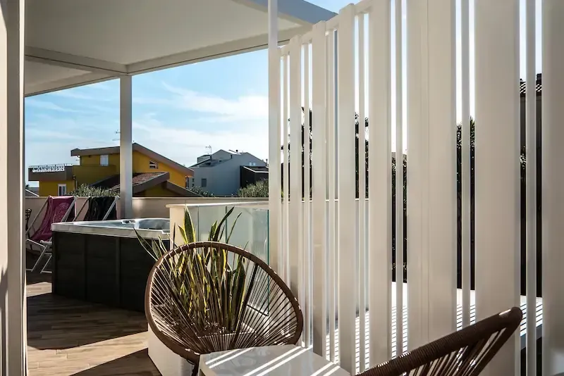 Balcony patio with wicker chair, white vertical blinds, and a view of houses under a blue sky