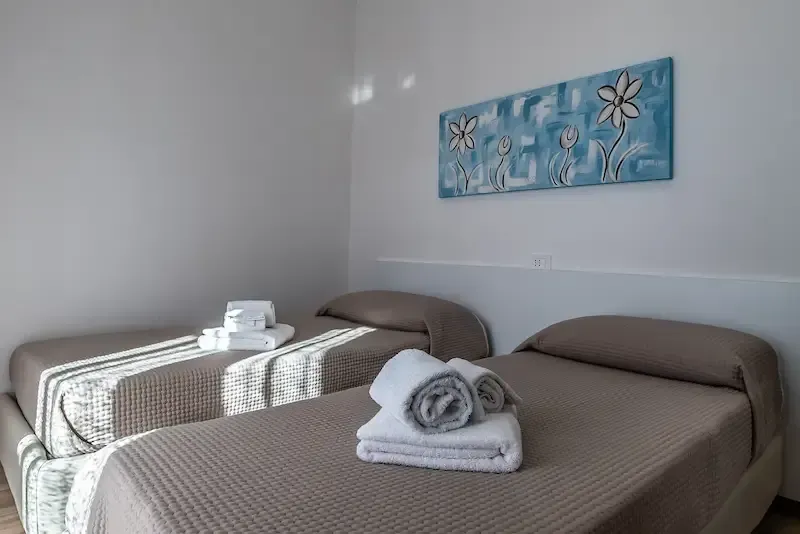 Two neatly made single beds in a bright room, with folded towels and a blue floral wall art piece