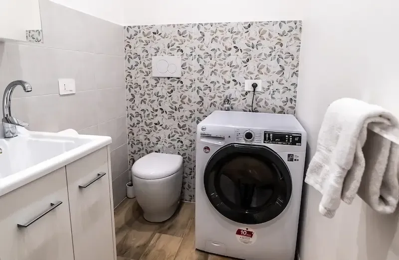 Compact bathroom with sink, toilet, and washing machine; floral backsplash and a towel hanging on the right.
