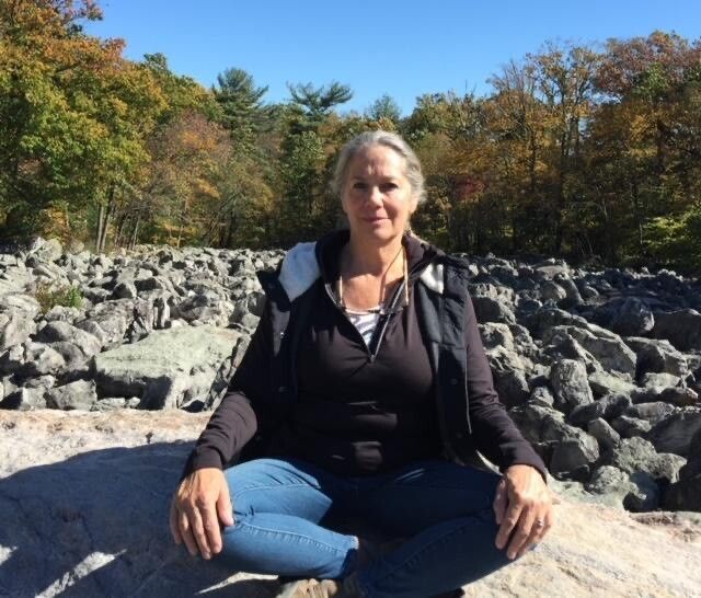 Woman sitting cross-legged on rocks outdoors; fall foliage in background.