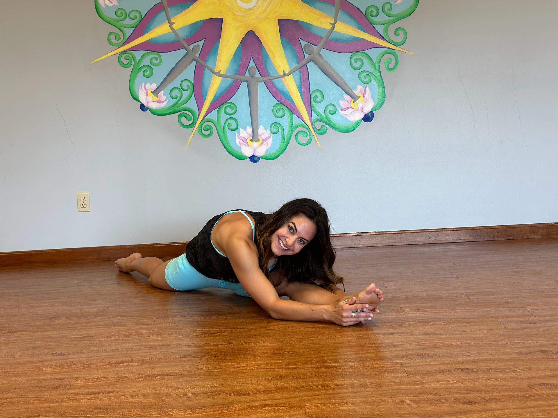 A woman is stretching on the floor in front of a painting on the wall.
