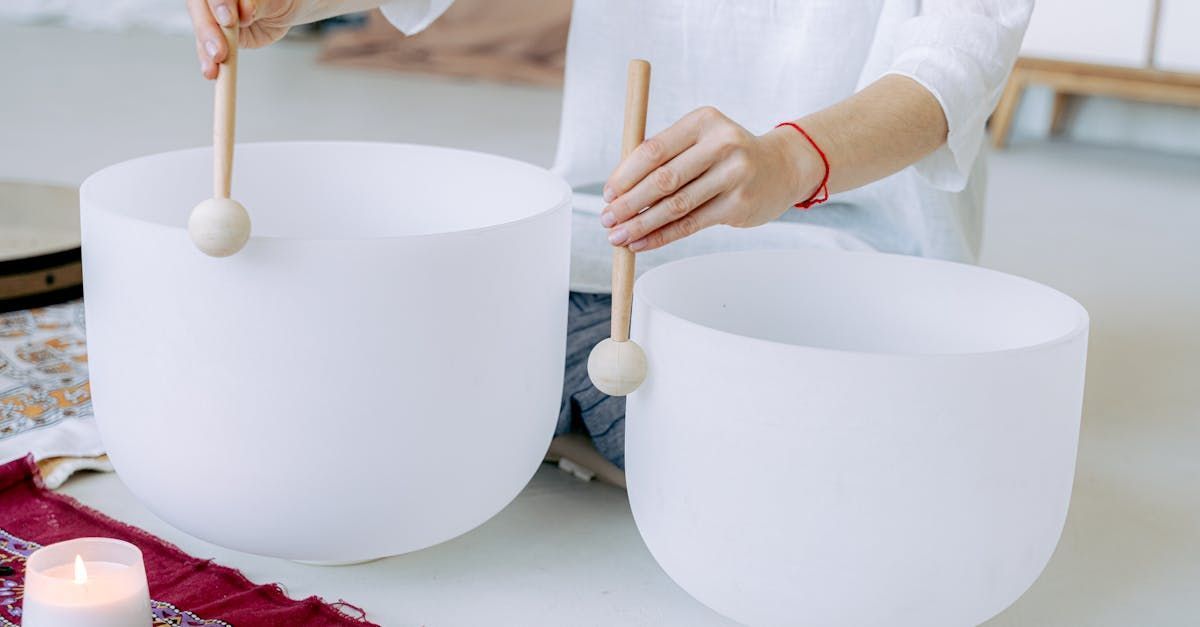 Person plays two large white crystal singing bowls with mallets. A candle burns nearby.