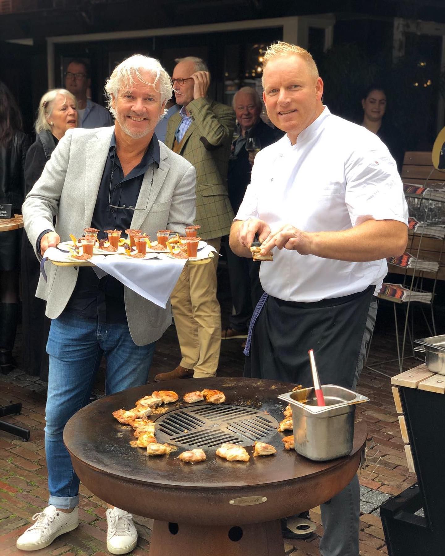 Twee mannen staan โโvoor een grill met schalen vol eten.