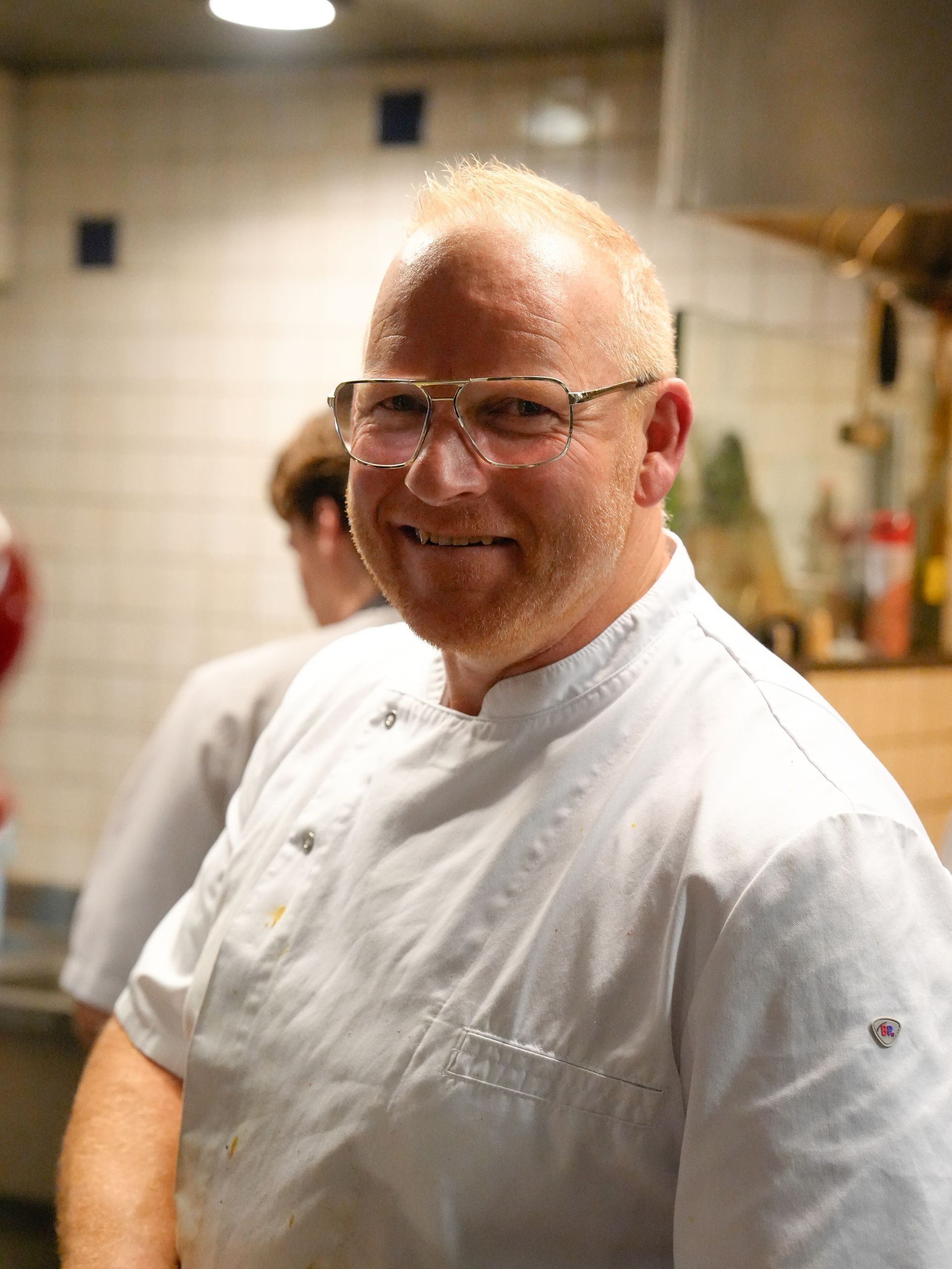 Een man met een bril en een wit overhemd lacht in een keuken.. Bij Linke Loetje