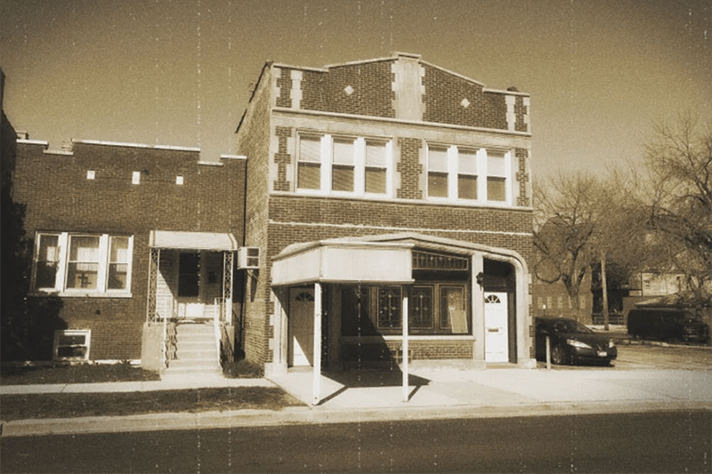 Sepia photo of two-story storefront buildings on a quiet street, with parked cars and bare trees