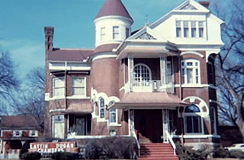 Large Victorian brick house with a turret and front porch, behind a hedge on a sunny day