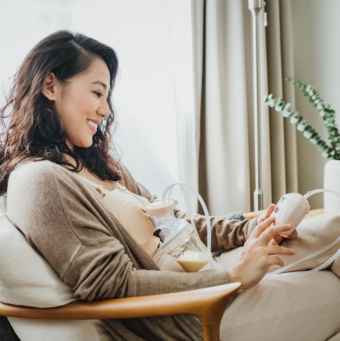 Woman using a breast pump, smiling while sitting in a chair indoors near a window.