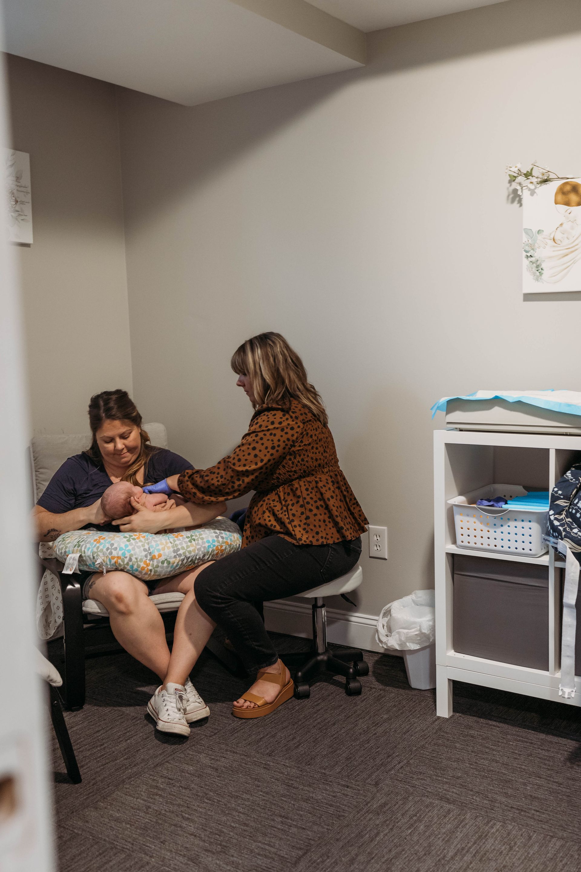A woman cradles a newborn while a lactation consultant assists. They're in a light-filled room.