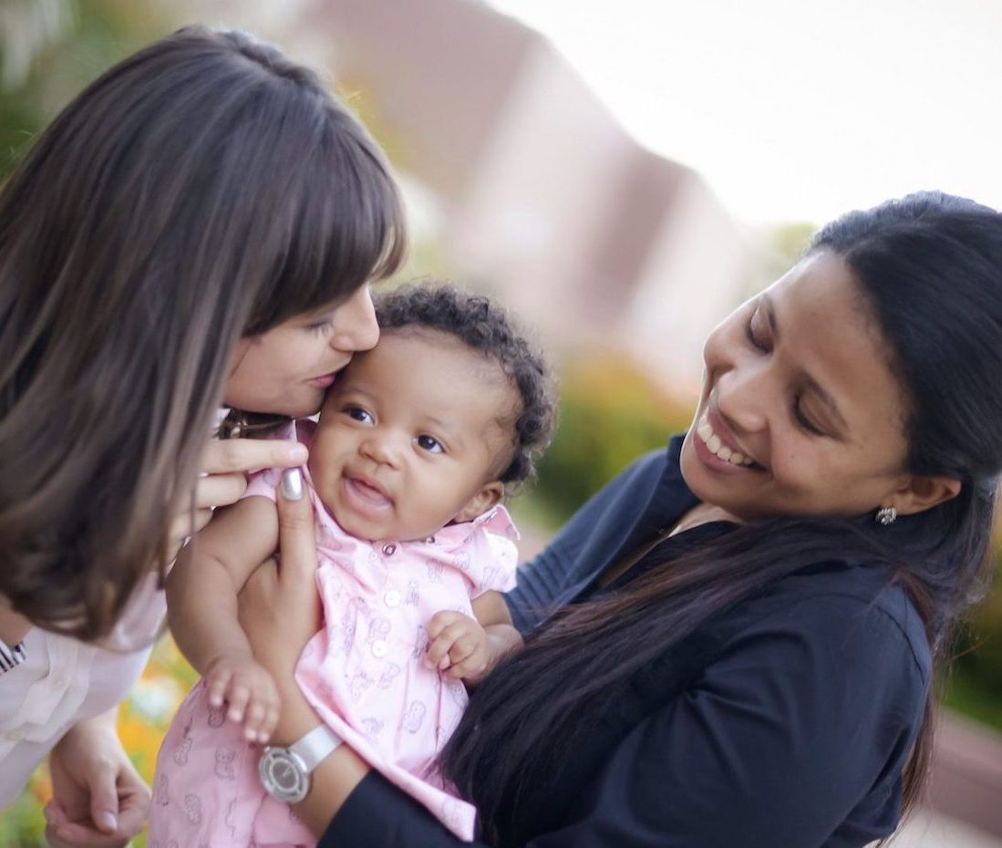 Two women smiling at baby wearing pink dress; one kisses the baby's cheek.
