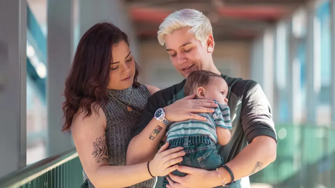 Two people holding a baby outdoors. One person has short, blonde hair; the other has dark hair. All are looking at the baby.