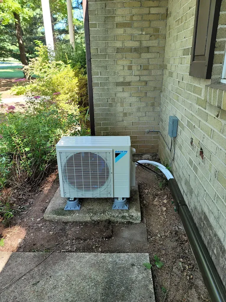 Outdoor air conditioning unit beside a brick house, with a concrete walkway and garden beds nearby